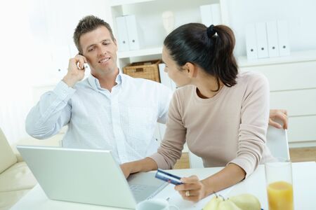 Young couple sitting at desk shopping on internet from home. Calling phone, using laptopcomputer and paying with credit card.の写真素材