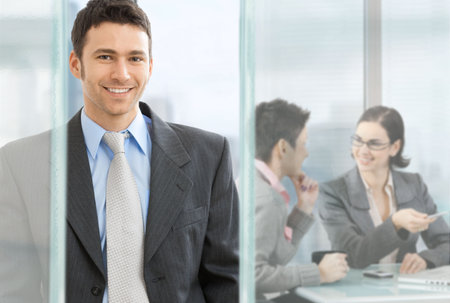 Happy businessman standing in modern glass office, businesswomen sitting at desk in the background,...