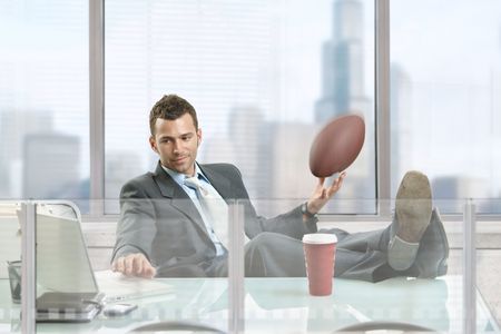 Relaxed businessman sitting at desk in front of office windows, holding football and smiling.の写真素材