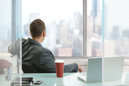 Relaxed businessman sitting at desk in office, looking out the windows to downtown skyscrapers.の写真素材