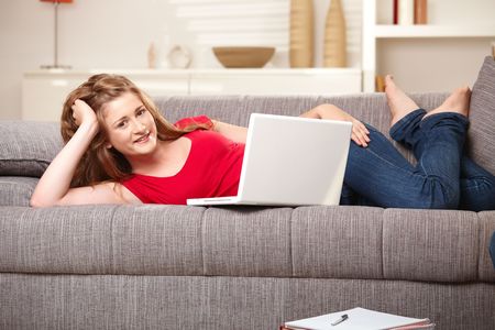 Smiling teen girl with laptop computer resting on sofa at home browsing internet looking at camera.の写真素材