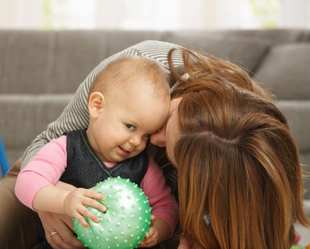 Baby girl smiling holding ball in hands, mum cuddling kissing baby on cheek.の写真素材
