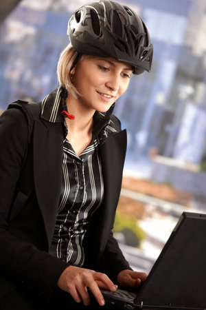 Portrait of young businesswoman wearing bike helmet, sitting in front of office building, using laptop computer.の写真素材
