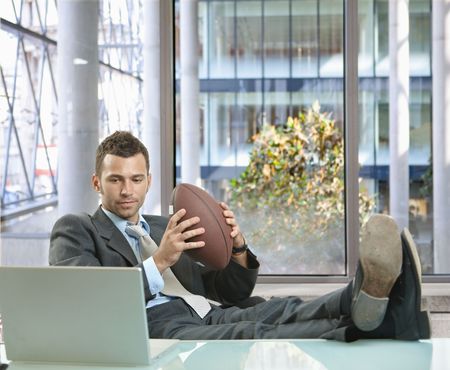 Relaxed businessman sitting at desk in front of office windows, holding football and smiling.の写真素材