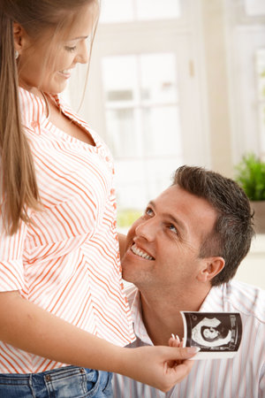 Happy man looking up at pregnant woman holding baby's ultrasound picture,smiling. 