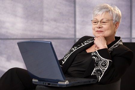Senior businesswoman sitting in office armchair, using laptop computer, smiling.の写真素材