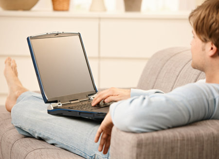 Man using laptop computer sitting on sofa in living room with feet up.