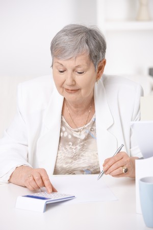 Senior woman sitting at desk using calculator.の写真素材