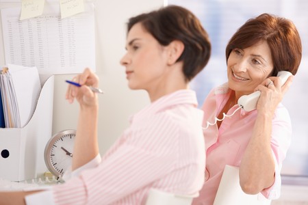 Female colleagues working together in office, mid-adult office worker holding pen, senior woman on landline phone.の写真素材