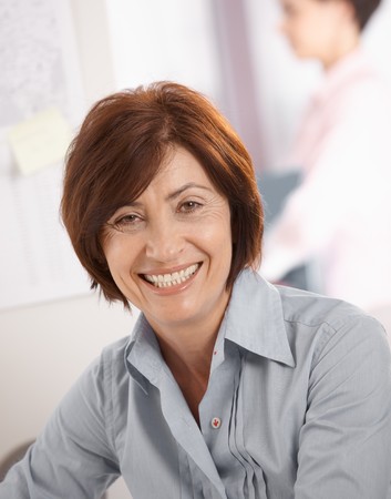 Portrait of senior businesswoman smiling at camera, coworker in background. Portrait of senior businesswoman smiling at camera, coworker in background.