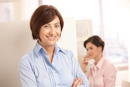 Senior professional woman smiling at camera, assistant using landline phone in background.の写真素材