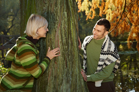 Young couple having fun in park, trying to catch each other around tree trunk at lake.の写真素材