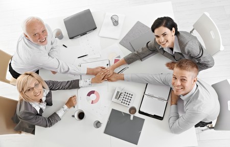 Confident businessteam holding hands at meeting over table expressing teamwork and unity, smiling at camera, overhead view.の写真素材