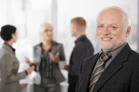 Portrait of senior businessman smiling at camera, team discussing work in background.の写真素材