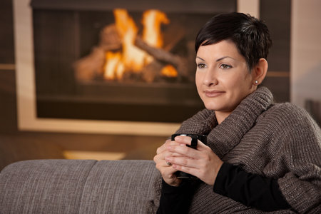 Woman sitting on sofa at home on a cold winter day, drinking hot tea, looking away.の写真素材