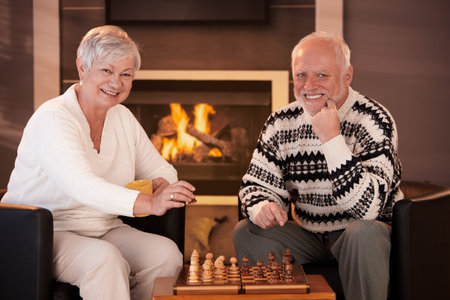 Portrait of elderly couple playing chess at home by fireplace in winter, looking at camera, smiling.の写真素材