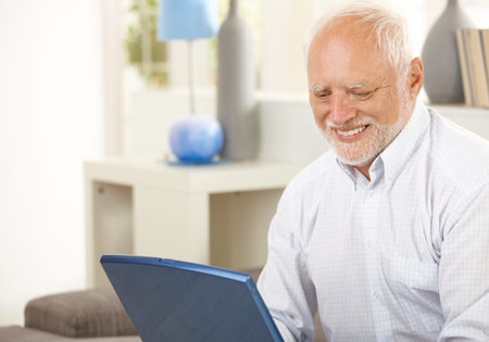 Portrait of aged man at home looking at laptop computer screen, smiling. 