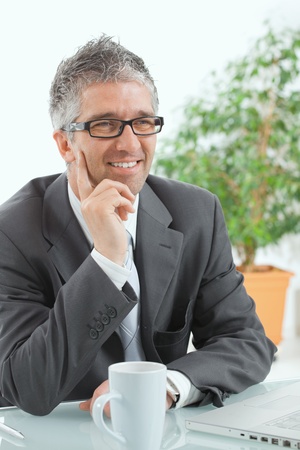 Businessman with grey hair, wearing grey suit and glasses thinking over laptop computer, sitting at desk, smiling.の写真素材