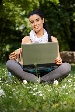 Pretty girl sitting in park with laptop and sandwitch, smiling, looking at camera.の写真素材