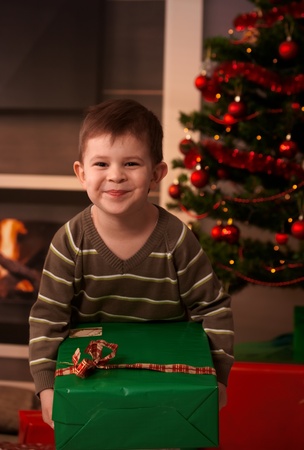 Happy kid sitting under christmas tree holding big wrapped box, laughing.の写真素材