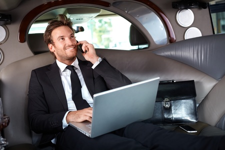 Smiling handsome businessman sitting in luxury limousine, working on laptop computer, smiling.の写真素材