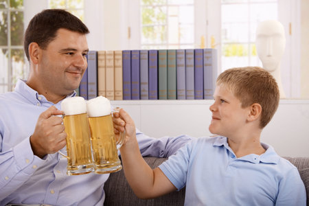 Father and son drinking beer, clinking with glasses, smiling.の写真素材