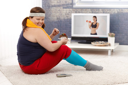 Overweight woman sitting on floor watching fitness program on television and eating chocolate cake,...