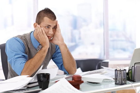 Troubled businessman sitting at desk covered with documents, chin in hand, thinking of problems. 