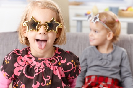 Little blond girl having fun at home, sticking tongue, wearing funny star shaped glasses, little sister in the background.の写真素材