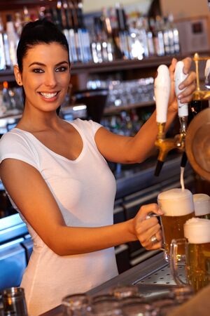 Portrait of attractive female bartender tapping beer in pub, looking at camera, smiling.の写真素材