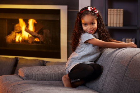 Beautiful afro little girl squatting on sofa in living room, smiling impishly.の写真素材