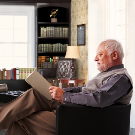 Elderly man sitting in armchair reading book at study at home.の写真素材