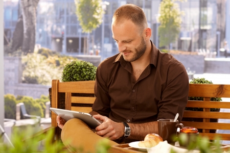 Casual man sitting in outdoor cafe on a bench, using tablet computer.の写真素材
