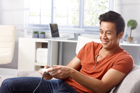 Young Asian man sitting in armchair at home, playing video game, smiing.の写真素材