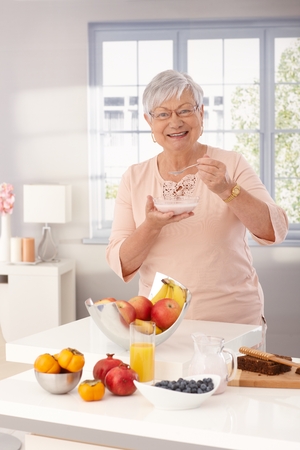 Happy grandmother eating breakfast cereal by kitchen counter full of fruits.の写真素材