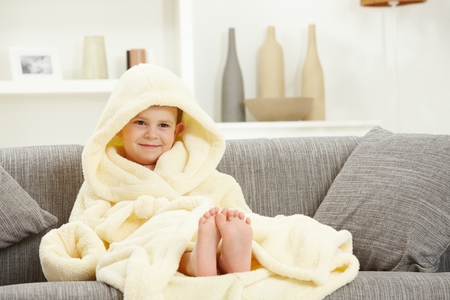 Happy smiling caucasian kid sitting in oversized bathrobe at home sofa, bare feet. Indoor, relaxed. Hood over head.の写真素材