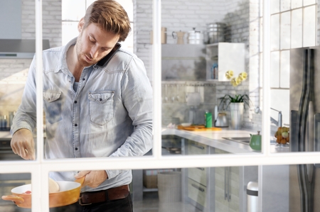 Casual young man cooking in kitchen chatting on phone, through the window view.の写真素材