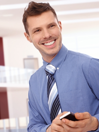 Cheerful young businessman with mobile phone at office center. Toothy smile, standing, tie, no jacket.の写真素材