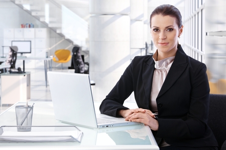 Attractive mid adult brunette caucasian businesswoman sitting at table with laptop computer at bright high-tech business center office. Smiling, looking at camera, woman suit.の写真素材