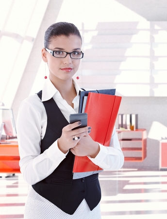 Casual young caucasian female secretary wearing glasses standing with folders and mobile phone in hand at bright office. Looking at camera, copyspace.の写真素材