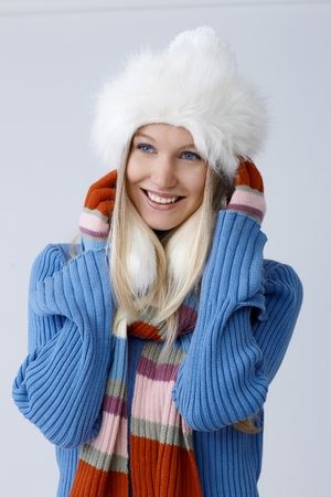 Portrait of young blonde woman in hat and scarf, smiling, framing face by hands, looking away.の写真素材