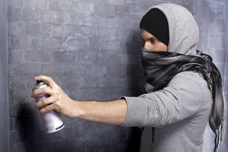 Graffiti man in hooded shirt and face mask spraying brick wall by aerosol can.の写真素材