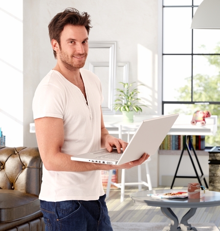Handsome young man standing in living room at home, holding laptop computer, smiling.の写真素材