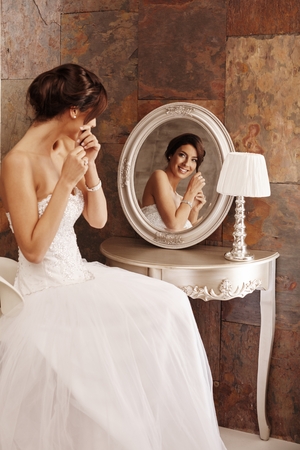 Beautiful young bride fixing knot of hair in mirror, smiling happy.の写真素材
