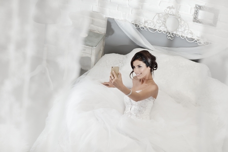 Happy bride making selfie by mobilephone while lying on bed in white bedroom.の写真素材