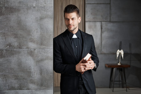 Portrait of good-looking young catholic priest holding book smiling. 