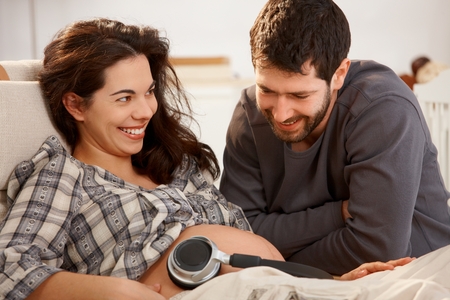 Happy pregnant woman resting in armchair at home husband holding earphones to naked belly.の写真素材