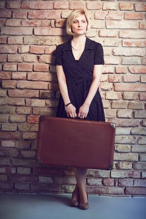 Old-fashioned photo of young woman standing against brick wall, holding suitcase.の写真素材