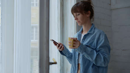 Woman at home at morning checking phone drinking coffee in windowの写真素材