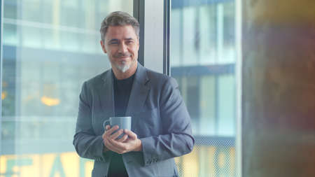 Happy older businessman taking break in office drinking coffee standing in window and daydreaming, thinking.の写真素材